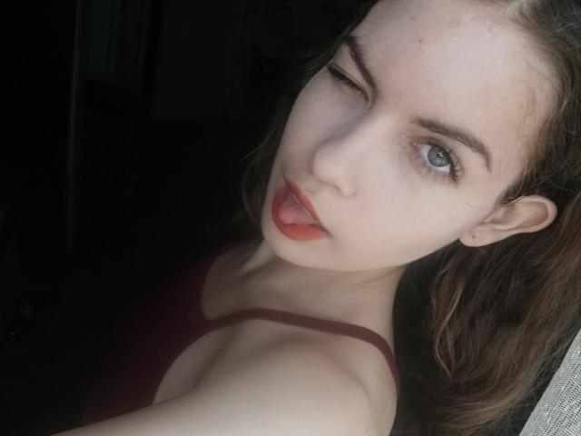 BrettRamsaroop with winking blue eye, tongue out, red lipstick, light skin, brown hair, wearing dark red bra, showing small tits, and a cute face in a selfie.