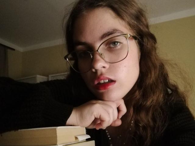 Brett Ramsaroop mit welligem, braunem Haar, Brille und rotem Lippenstift, mit dem Kinn in der Hand, wirkt leicht verärgert, trägt ein schwarzes Oberteil, im Vordergrund befinden sich Bücher..