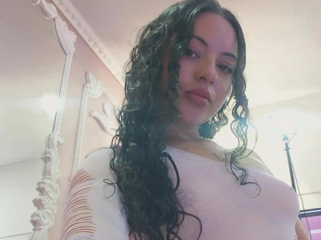 MiiaBennet, avec ses cheveux noirs bouclés, portant un haut blanc transparent qui révèle ses petites seins, et affichant une expression neutre, se tient debout dans une pièce éclairée doucement..