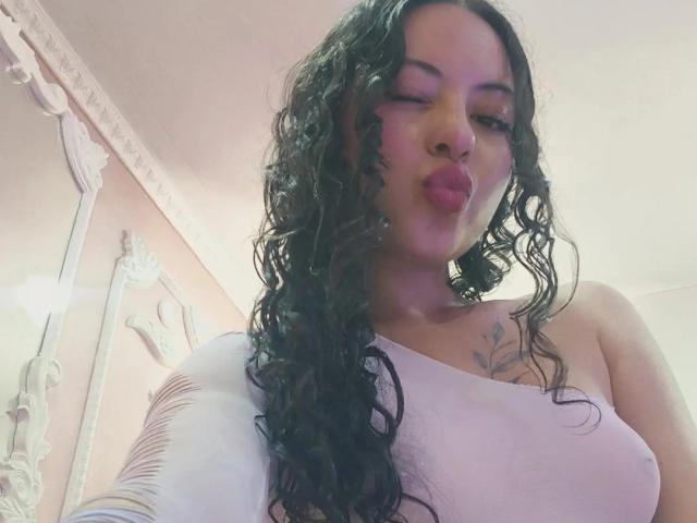 MiiaBennet, com cabelo preto encaracolado, lábios rosados e finos, uma blusa branca de um ombro só e uma pequena tatuagem no peito direito, olhando para baixo para a câmera. Seios visíveis..