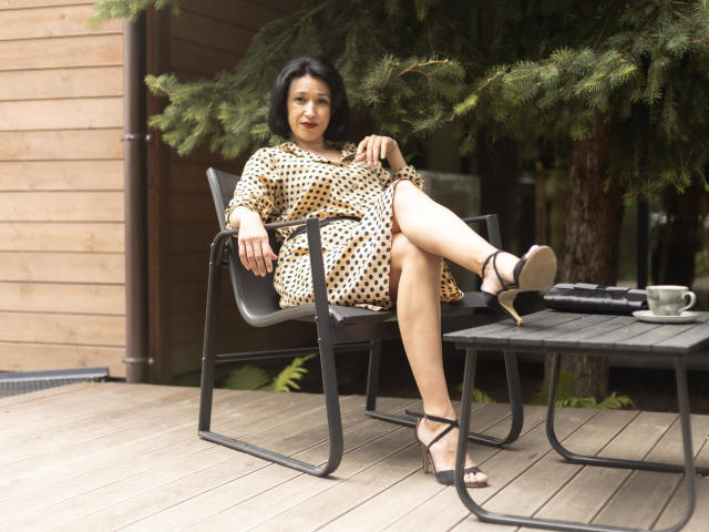 CameronEnns sits on a black chair, legs crossed, wearing a polka dot dress, black strappy heels, with a relaxed smile, in a wooden outdoor setting, by a green tree.