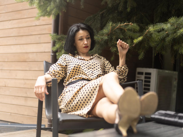 CameronEnns, middle-aged woman with black bob, wearing polka dot dress, sitting on black chair, holding pine branch, legs crossed, beige heels, wooden wall, slightly annoyed expression.