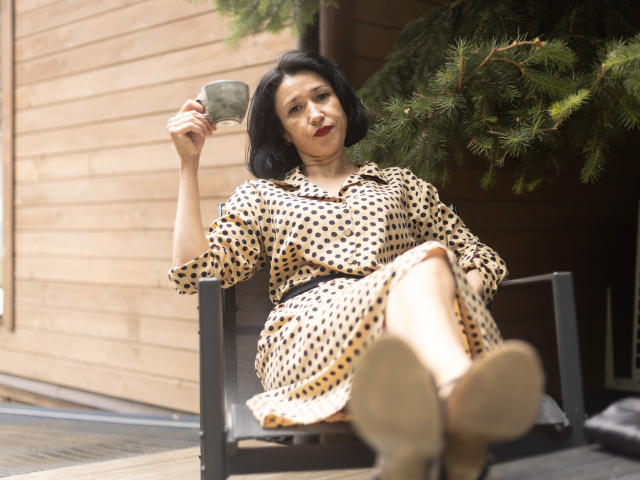 CameronEnns with short black hair, polka dot dress, holding a green mug, sitting on a chair, legs crossed, looking up, green tree branch in background, casual, relaxed.
