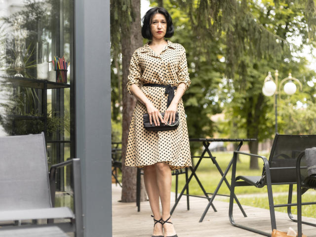 Cameron Enns steht draußen, trägt ein beiges Kleid mit schwarzen Punkten und einen schwarzen Gürtel, hält eine schwarze Clutch, trägt beige High Heels, hat kurze schwarze Haare und einen ernsten Gesichtsausdruck..