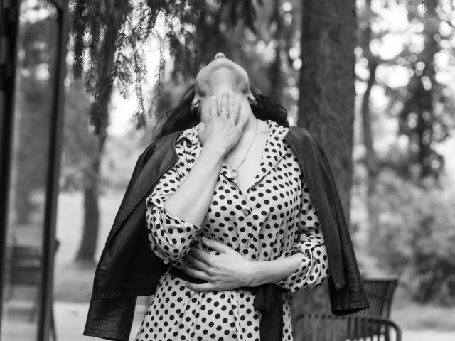 CameronEnns with polka-dot dress, leather jacket, head tilted back, hand covering mouth, other hand on belly, fingers showing a ring, black-and-white photo, tree in background.