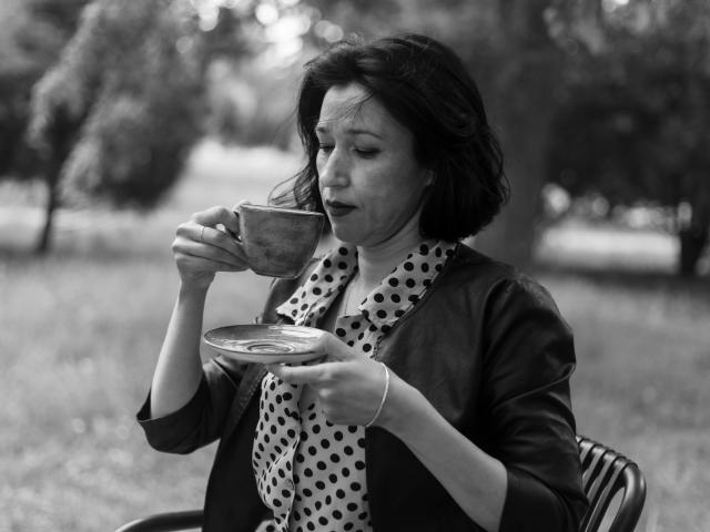 Schwarz-weißes Foto von CameronEnns mit kurzen, dunklen Haaren, die aus einer Keramiktasse trinken, einen Untertasse halten, einen Punkte-Schal und eine dunkle Jacke tragen und leicht verärgert aussehen..