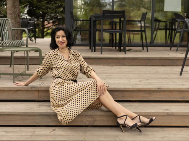 CameronEnns, sitting on wooden steps, wearing a polka dot dress, black belt, and black heels, with short black hair, red lipstick, and a smile, legs crossed.