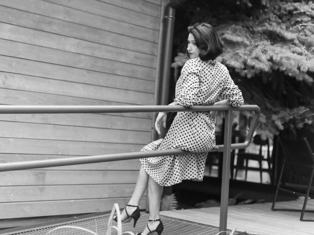 CameronEnns est assise sur une rambarde en métal, vêtue d'une robe à pois et de talons hauts, avec ses fesses légèrement relevées, regardant sur le côté, sur une photo en noir et blanc..