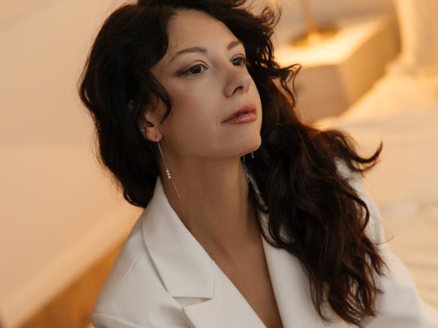 PeachMirage with wavy dark brown hair, wearing a white coat, looking slightly up with subtle makeup and small earrings, in a softly lit, warm-toned room.