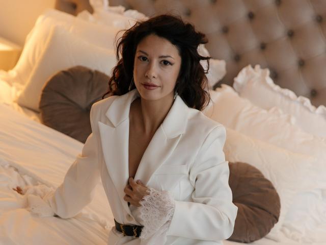 PeachMirage with curly dark hair, wearing a white blazer, lace cuff, and belt, sitting on a bed with white pillows and brown cushions, looking at the camera.