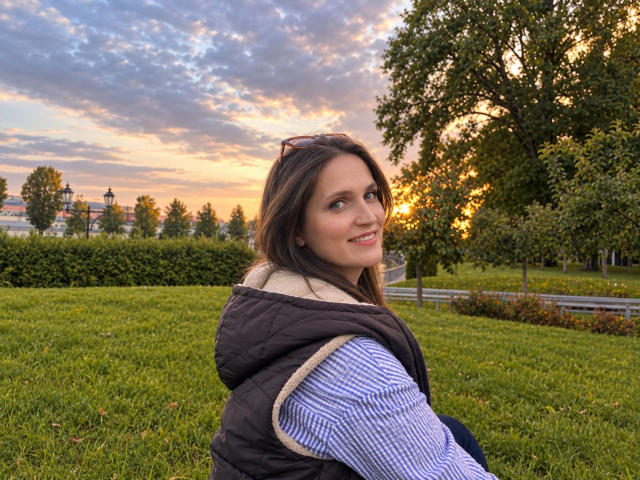 LoraParkerHot has tits, brown hair, and a cute smile, wearing a black puffy vest over a blue-striped shirt, sitting on green grass at sunset with a tree behind.