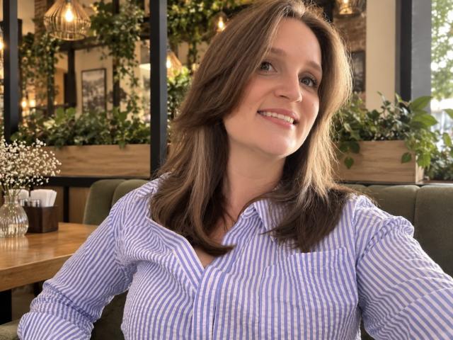 LoraParker: Una mujer de cabello castaño, sonriendo, vestida con una camisa a rayas moradas y blancas, sentada en un asiento verde, con una mesa de madera y flores, y luces cálidas en el fondo..