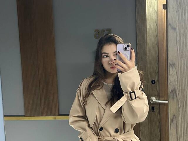 Anne Kofron, avec de longs cheveux bruns, portant un trench beige, tenant un iPhone rose, prenant un selfie devant un miroir, montrant son visage et le bouton noir de son manteau..