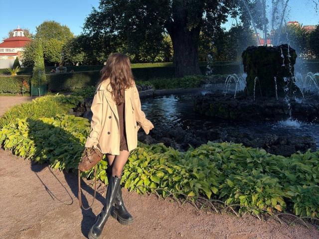 AnneKofron staat bij een fontein, gekleed in een beige jas, zwarte laarzen en een bruine jurk, met lang bruin haar, en houdt een bruine tas vast, in een zonnige tuin..