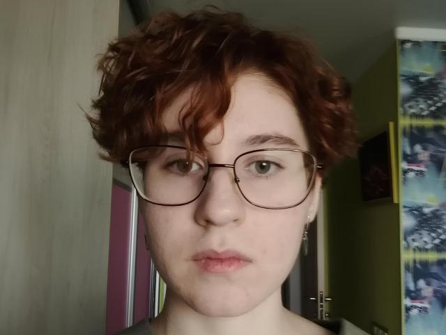 Lourie Morphew hat lockiges, rotes Haar, eine Brille, blasse Haut und wirkt ernst, mit leicht geöffneten, rosafarbenen Lippen. Ihre grünen Augen sind im Bild zentral platziert..
