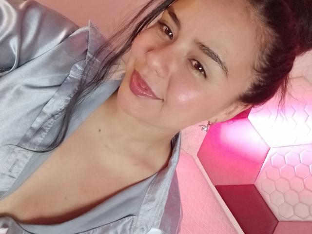 ArielleWilde with shiny pink lips, dark hair in a ponytail, wearing a silky gray shirt, showing a cute smile, and small tits, in a pink-lit room.