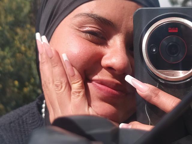 ArielleWilde with tan skin, black headscarf, and long white nails, smiling while holding a camera to her face, showing off her cute nose and small, slightly upturned lips.