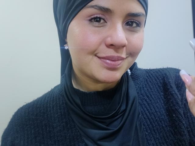 Arielle Wilde, con un hijab nero e un maglione a maglia nero, ha un sorriso carino e occhi marroni, mostrando il suo naso e le labbra rosa, mentre tiene una sigaretta nella mano destra..