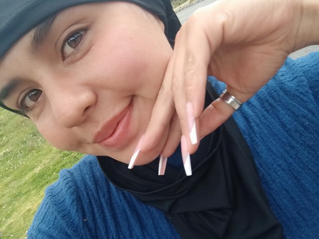 ArielleWilde's face, smiling with a hand touching her cheek, has long white nails and a silver ring on her finger. She's wearing a blue knit sweater and black headscarf.