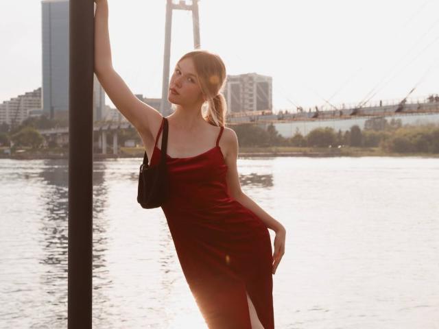 MicheleIreland with red dress, black purse, and blonde ponytail, leaning on pole, sunlight, water, city buildings, and bridge in background, her tits slightly visible, sexy, elegant.