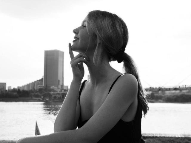 Michele Ireland, les lèvres pincées, un doigt sur le menton, un débardeur noir, une queue de cheval, vue de profil, montrant légèrement ses seins, en noir et blanc, près d'un plan d'eau avec un grand bâtiment..