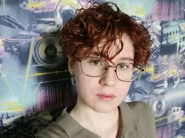 LourieMorphew heeft krullend rood haar, een bril, bleke huid en een neutrale uitdrukking, en draagt een grijze trui. Hun kleine borsten zijn subtiel zichtbaar. De foto is een selfie..