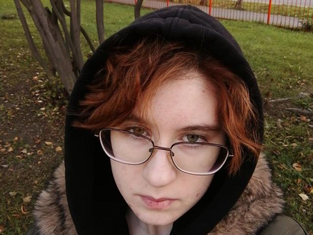 LourieMorphew hat rotes, zerzaustes Haar, eine schwarze Kapuze und eine Brille mit einem ernsten Gesichtsausdruck. Sie trägt eine mit Fell gefütterte Jacke und befindet sich draußen in einer grasbewachsenen Gegend..