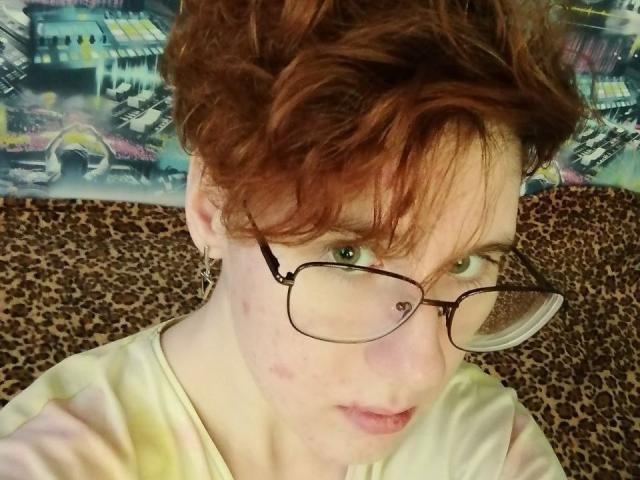 LourieMorphew has curly red hair, green eyes, glasses, pale skin, small zit on cheek, and a leopard-print shirt, looking at camera with a slightly pouty expression.