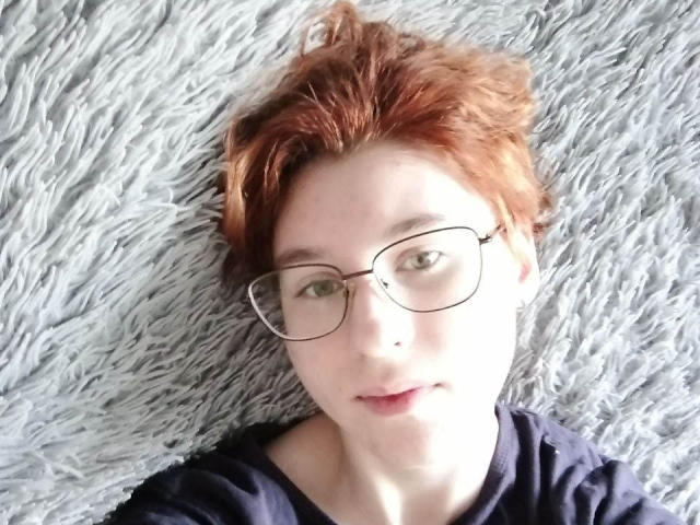 Lourie Morphew, con cabello rojo, gafas y piel pálida, acostada sobre una alfombra gris y esponjosa, vestida con una camisa oscura, mirando a la cámara con una ligera sonrisa..