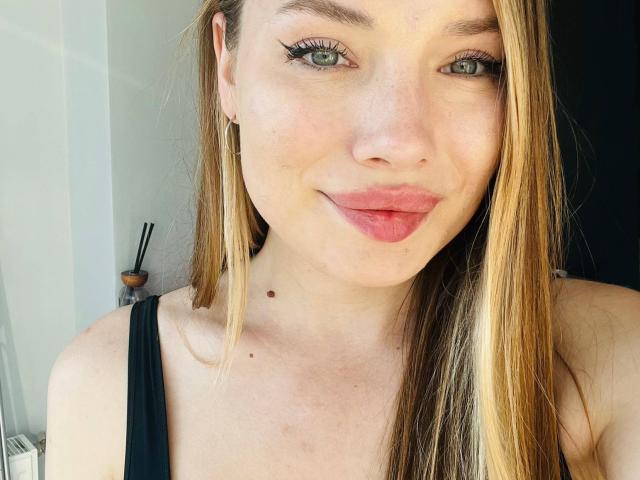 IceDaniellee has light green eyes, long blonde hair, pink lipstick, and black eyeliner, wearing a black tank top with small tits and a cute mole on her chest.