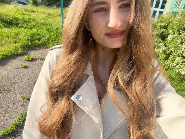 MontanaX has long, wavy brown hair, wearing a beige jacket, with small tits and a cute smile, standing in a sunny grassy area, looking at the camera.