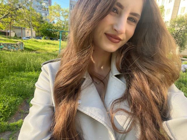 MontanaX with long brown wavy hair, wearing a white jacket, smiling, showing a cute face, brown lipstick, and a necklace with a small pendant, in a sunny green park.