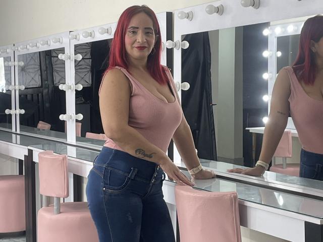 KateMonroe with red hair, pink tank top, and blue jeans, standing in a mirror-lined room, showing her tits and a cursive tattoo on her right forearm.
