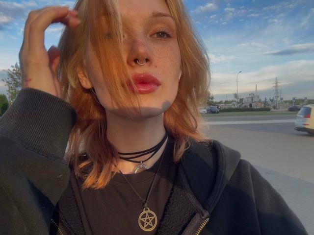 Aline Gossom, con i capelli rossi, un anello al naso e una collana nera, indossa una collana con un pentagramma, ha le labbra piene, un cappuccio nero, una mano tra i capelli e un cielo blu sullo sfondo..