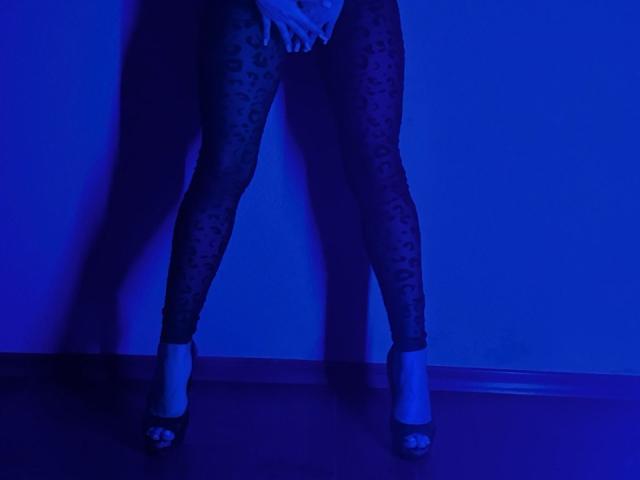 Les jambes de BalletSwan sont habillées de collants noirs à imprimé léopard transparent. Elle se tient debout, en talons hauts, les mains couvrant ses parties intimes, sous un éclairage bleu..