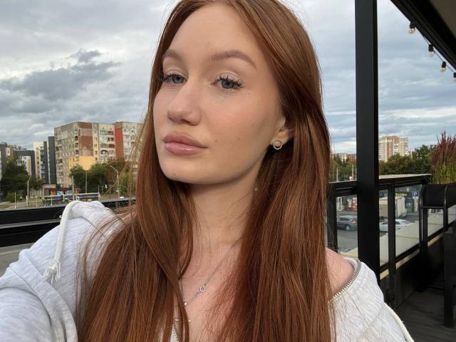 CamillaRosee with long brown hair, light skin, subtle makeup, small earrings, white hoodie, pink lips, and a confident expression, standing outside a glass building.