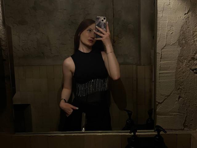 CamillaRosee with long brown hair, wearing a black sleeveless dress with silver fringe, taking a mirror selfie in a dimly lit, textured room. Her hand points at her chest.
