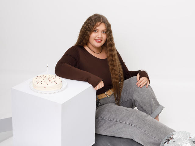 Madrinia, avec de longs cheveux bruns bouclés, un haut marron, un pantalon gris et une ceinture beige, assise sur un sol blanc, un bras posé sur un cube blanc, un gâteau blanc avec des vermicelles rouges sur le dessus..