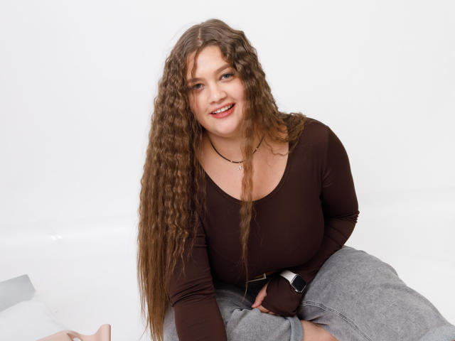 Madrinia a de longs cheveux bruns bouclés, porte un haut marron à manches longues et un pantalon gris, elle sourit et porte un collier noir ainsi qu'une montre, assise sur une surface blanche..