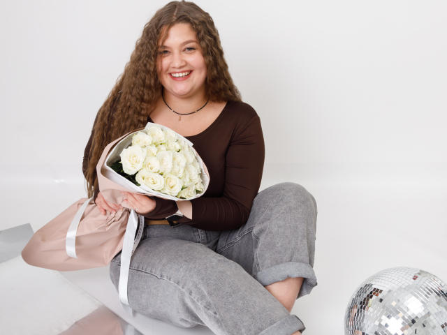 Eine Frau namens Madrinia mit lockigem, braunem Haar, die lächelt und ein braunes Oberteil mit langen Ärmeln und eine graue Hose trägt. Sie hält einen Strauß weißer Rosen, sitzt mit einer pinken Tasche und einer silbernen Discokugel..