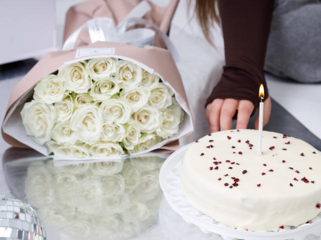 Madrinias Hand berührt eine weiße Torte mit roten Beeren und einer Kerze, während ein Strauß weißer Rosen auf einer reflektierenden Oberfläche liegt..