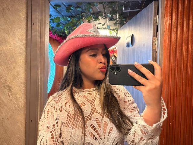 LiaDiane, con la pelle abbronzata, i lunghi capelli scuri, indossa un cappello da cowboy rosa e una camicetta bianca in pizzo, sta scattando un selfie allo specchio con la mano destra, mostrando le sue labbra rosse..