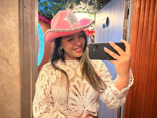 LiaDiane mit rosa Cowboyhut und weißem Spitzenoberteil, hält ein Telefon, macht ein Selfie im Spiegel und zeigt ihre Brüste durch die Spitze, lächelt mit herausgesteckter Zunge..