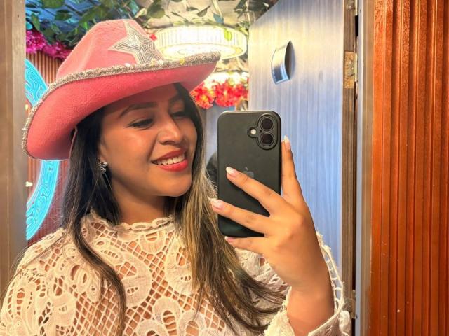 LiaDiane with brown skin, long hair, smiling, wearing a pink cowboy hat with a star and white lace top, taking a mirror selfie with a black phone.