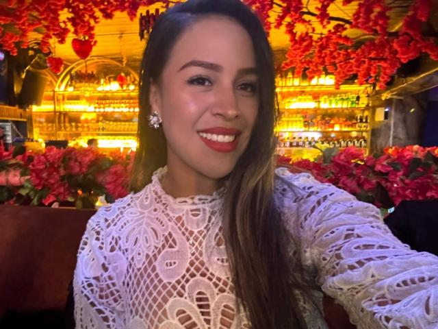 LiaDiane met lang bruin haar, die een witte kanten top draagt, lachend, met rode lipstick, oorbellen, en die een selfie maakt. Borsten niet zichtbaar, focus op het gezicht en de top..