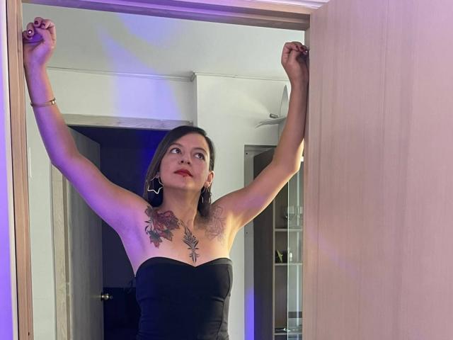 ArianaBlonde stands in a doorway with arms raised, showing off her tits and tattoos, wearing a black strapless dress, looking up with red lipstick and earrings.