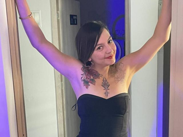 ArianaBlonde with arms raised, black strapless top, red lipstick, tattoos on chest and arm, smiling, brown hair, standing in doorway, purple and blue lighting.