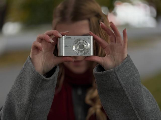 Norah Habrock segurando uma câmera Sony com as duas mãos, o rosto parcialmente obscurecido, vestindo um suéter cinza e um cachecol vermelho, fundo desfocado..