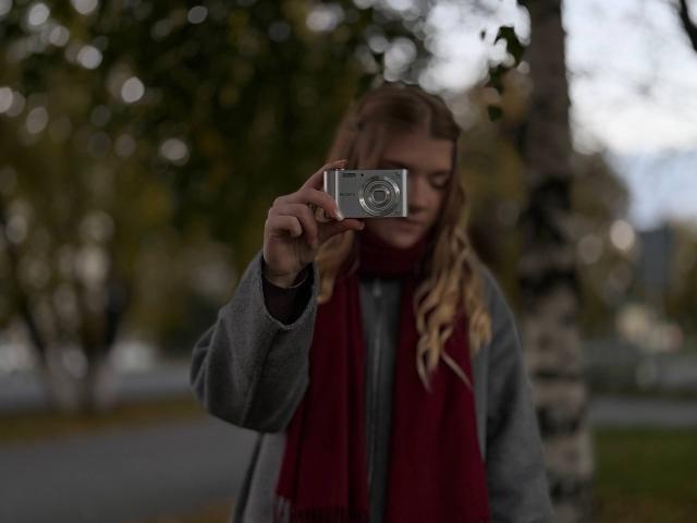 Norah Habrock, con i capelli biondi, una sciarpa rossa e un cappotto grigio, tiene una macchina fotografica davanti al viso, scattando una foto. Lo sfondo è sfocato, l'ambientazione è autunnale, la luce è soffusa e l'attenzione è focalizzata sulla macchina fotografica..