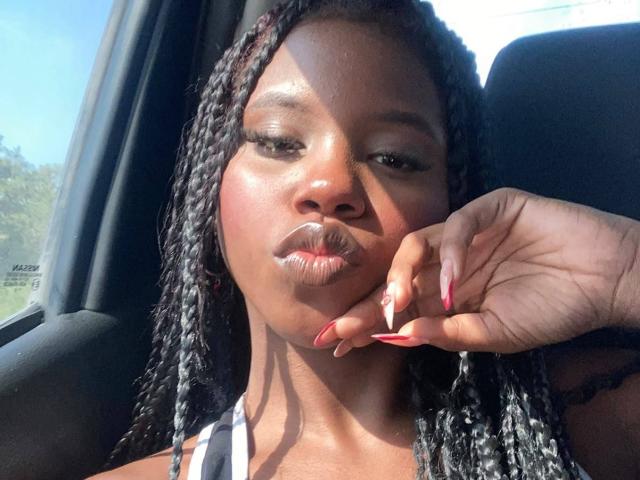 CatalinaSanA with dark skin and braided hair, pouting lips, red nails, and a hand resting on her cheek in a car. Sunlight highlights her glossy skin.